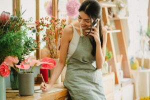 An image of a woman talking on the phone happily while leaning on her desk, writing in her notebook at the same time.