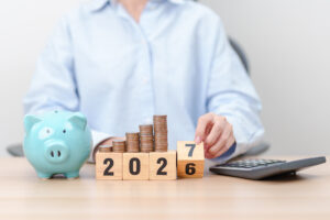 Piggy bank, rising stacks of coins and year blocks changing from 2026 to 2027 beside a calculator.