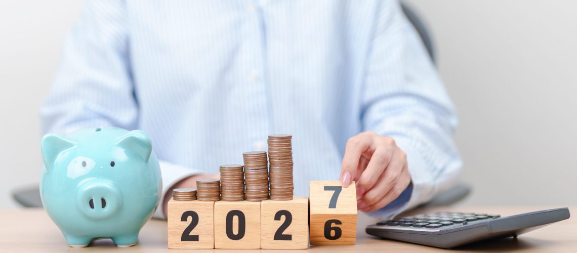 Piggy bank, rising stacks of coins and year blocks changing from 2026 to 2027 beside a calculator.