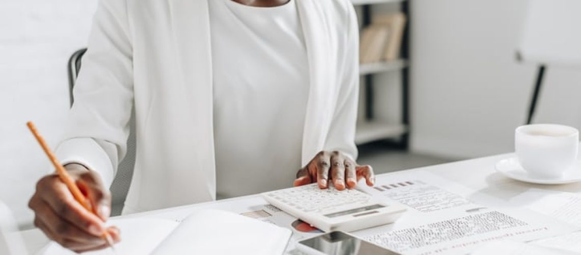 cropped view of african american adult businesswoman in white formal wear writing in notebook and working at office desk