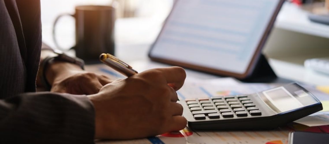 Millennial businessman working with digital tablet, using calculator to check the statistics and projections.