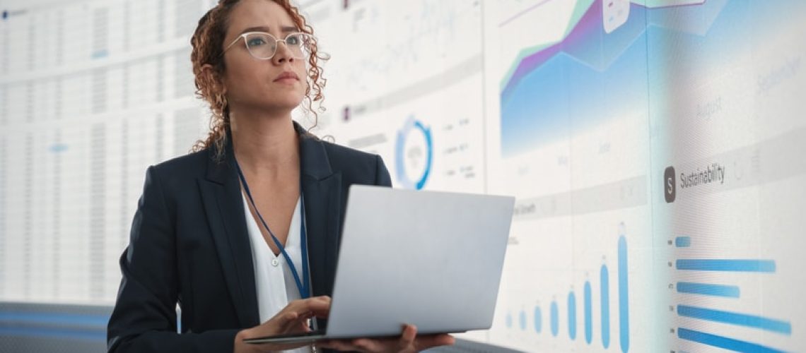 In the Monitoring Office: Successful Hispanic Businesswoman Holding Laptop Computer And Analyzing Data On Big Digital Screen With Graphs and Charts. Female IT Entrepreneur Running Software Company.