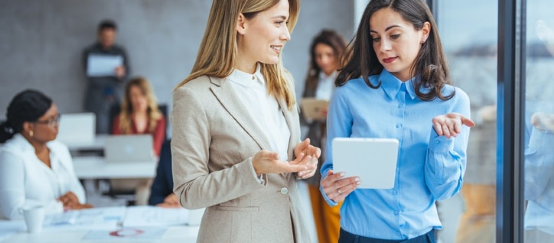 Two professional women in smart attire are actively discussing a project on a tablet, with a diverse group of colleagues working in the background.